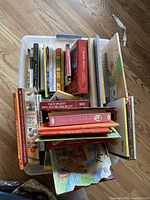 Top-down view of a plastic tub filled with assorted books showing various colorful covers and spines, some titles visible including 'Cousin Bobby' and several children's books.
