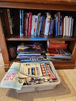 Bookshelves with assorted books about New York, Broadway, and theater, and a plastic bin of magazines on the floor.