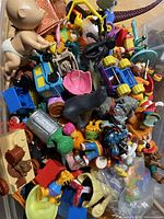 Photo showing a pile of assorted small plastic figurines and toys including characters from Garfield, Smurfs, Muppets, along with animal and vehicle toys such as a black cat, large spider, and dinosaur tail figure.