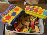 Bin filled with various wooden blocks and colorful plastic musical toy pieces including a yellow toy piano with colored keys.