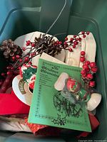 Photo showing lot contents including artificial pine cones, red berries, holiday ribbon, a small ceramic figurine with Christmas bells, and a Christmas-themed book.
