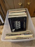 Clear plastic bin holding a large collection of vinyl LP records, showing the Star Wars album cover on top in the first image.