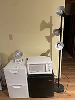 Photo showing mini fridges, microwave, floor lamp and fan, all placed against a wall on wood floor.