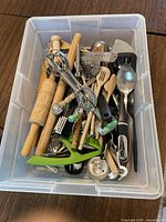 Photo shows a clear plastic storage bin filled with assorted cooking utensils including rolling pins, wooden spoons, metal spatulas, and a hand crank egg beater.