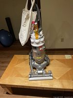 Full front view of the Dyson DC15 upright vacuum cleaner standing on a wooden table with a cloth tote hanging on the handle and the attached hose and wand visible.