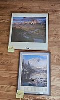 Full view of two framed mountain landscape photographs on a wooden floor: one of the Grand Tetons and one of the Maroon Bells with visible wooden and blue frames.