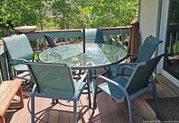Photo showing round glass top patio table with six blue mesh fabric chairs around it, on a wooden deck with an umbrella stand.