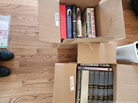Two cardboard boxes filled with various hardcover books on fiction and history, placed on hardwood floor.
