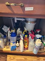 Photo of assorted used cleaning supplies stored under kitchen sink, showing diverse bottles and containers including Lysol spray, white pump bottle, and spray bottles with red triggers.