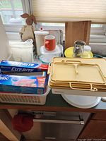 Photo showing plastic storage containers, boxes of aluminum foil and plastic bags, and various kitchen items including a plastic colander and small utensils.