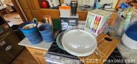 Kitchen items on counter including blue bowls, ceramic plate with glass dome lid, wooden cutting board, colorful small glasses box, black travel mug, and other glassware.
