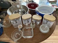 Overview of assorted kitchen items including metal mixing bowls stacked inside each other, clear plastic decorative containers with wooden lids on the table, blue and white plastic colander, red bundt cake pan, large clear glass bowl, and small glass dishes and bottles.