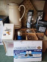 Photo showing white insulated pitcher, collectible Tom and Jerry figurine box, assorted stemmed wine glasses, and various water and air filters including LG brand and charcoal filters.