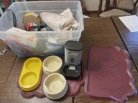 Photo shows a large clear plastic tub filled with assorted pet supplies on a table alongside pet bowls, an automatic feeder, and a paw print silicone mat.