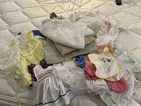 Multiple assorted baby clothes laid out on a mattress showing dresses, knitted items, bonnet, bib, and other pieces.
