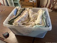 Plastic bin tightly packed with various folded blankets and sheets including leaf pattern fleece blanket and solid color linens