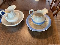 Two antique wash pitcher and bowl sets placed on a wooden table. One set is blue with floral decoration and the other is plain white. Both pitchers have large handles and scalloped rims.
