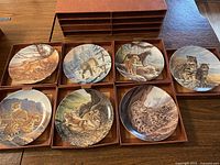 Lot of 8 Knowles big cats collector plates arranged in and beside a brown 8-drawer cardboard storage box. Plates depict various big cats such as lions, cougars, bobcats, cheetahs with artwork by Lee Cable.