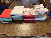Full view of the lot showing stacked reams of printer paper in white, red packaging, and assorted envelopes.