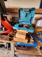 Photo showing a blue Makita carrying case containing a cordless drill, battery, and charger with orange extension cords coiled beside it on a wooden workbench.