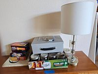 Photo of a grey metal lock box with key, clear glass table lamp with white shade, and four Ball Mason jars in box on wooden surface.