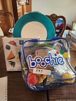 Clear plastic Boochie bag with assorted craft items inside, a round turquoise plate or frame, and a box of seashells with colorful origami starfish graphics.