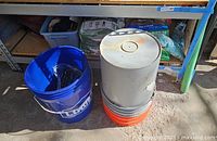 Photo shows two large bags of fertilizer and soil products (Scotts and Miracle Grow) on shelf, two bags of Quikrete concrete mix, three 5-gallon buckets (blue, orange, gray), and a plastic tote containing miscellaneous items.