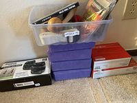 Clear view of plastic storage bin containing various health and fitness accessories including hand warmers, a wooden massage ball, and a white strap. Underneath, four purple foam yoga or exercise blocks stacked. To the side, boxed reusable heating pad packages in red boxes and a Reebok black ankle weights box.