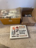Full overview of the lot including the yellow sewing box, cardboard box with yarn, and the sewing book spread on the carpeted floor.