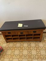 Front view of wooden mudroom bench with three drawers and shoe cubbies below, topped with a black faux leather cushioned seat.