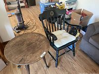 Photo showing small round wood side table with significant wear and black wooden armchair with University of Colorado Boulder seal on backrest and white seat cushion
