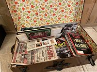 Open small brown vintage trunk showing interior floral lining and magazines and newspapers inside.