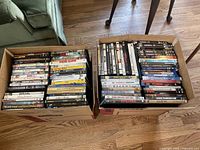 Two large cardboard boxes filled with DVD and Blu-ray cases, stacked on a wood floor next to a green sofa. Visible titles include Home Alone and Young Frankenstein.