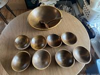 Top view showing one large wooden salad bowl, two wooden salad forks inside it, and eight smaller wooden serving bowls arranged on a round wooden table.