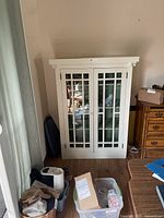 Full view of white wood storage cabinet with glass panel doors standing against a wall on wooden floor.