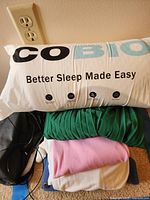 Better Sleep pillow on top of folded clothes including black jacket, green dress, pink top, and white or beige item.
