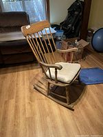 Wooden rocking chair seen from side angle showing structure and cushion on seat. The chair shows visible wear on wood finish.