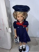 Front view of Shirley Temple doll in navy sailor dress and hat, holding shoes, standing on a support stand, next to box.