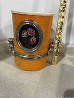 Full view of the orange vase planter showing size against a measuring stick, metal flower decoration on the front, and silver metallic bands around the body.
