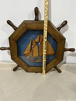 Front view showing the wood carved ship wheel frame and 3D schooner model inside with ruler for scale
