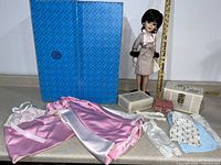 Full view of the doll standing next to a blue box with assorted accessories including two suitcase cases, pink satin nightgown, and other fabric items.