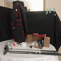 Golf set arranged showing golf bag, golf clubs, white ceramic mug, boxes of golf balls, and bags of golf tees on white cloth.