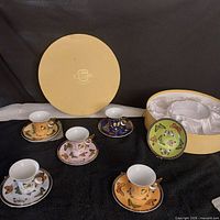 Five butterfly-themed espresso cups each with a different colored background and matching saucers, displayed alongside a round beige satin-lined box with Duchess logo.
