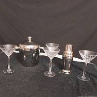 Four clear crystal martini glasses arranged with a metal ice bucket and a metal martini shaker all displayed against a black background.