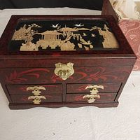 Front view of the cherry wood lacquered jewelry box with ornate cork art under glass lid and gold latch and handles on drawers.