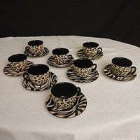 Eight hand-painted ceramic cups and saucers displayed on a white cloth, showing the animal print patterns and black interiors.