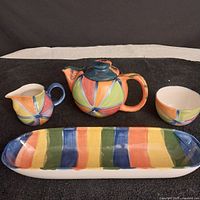 Front view photo of the teapot, creamer, sugar bowl and boat-shaped bowl with colorful vertical stripes in blue, green, orange, yellow, and pink.