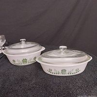 Two oval-shaped white opaque glass casserole dishes with green floral patterns side by side with clear glass lids on black background.