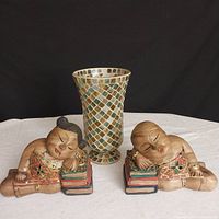 Pair of wooden carved and painted sleeping Buddha boy and girl book ends with colorful embellished details, placed on either side of a mosaic glass tile hurricane vase with gold, green, and blue tiles.
