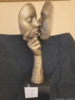 Front view of the bronze-tone resin sculpture showing two masks kissing and hand support on black base